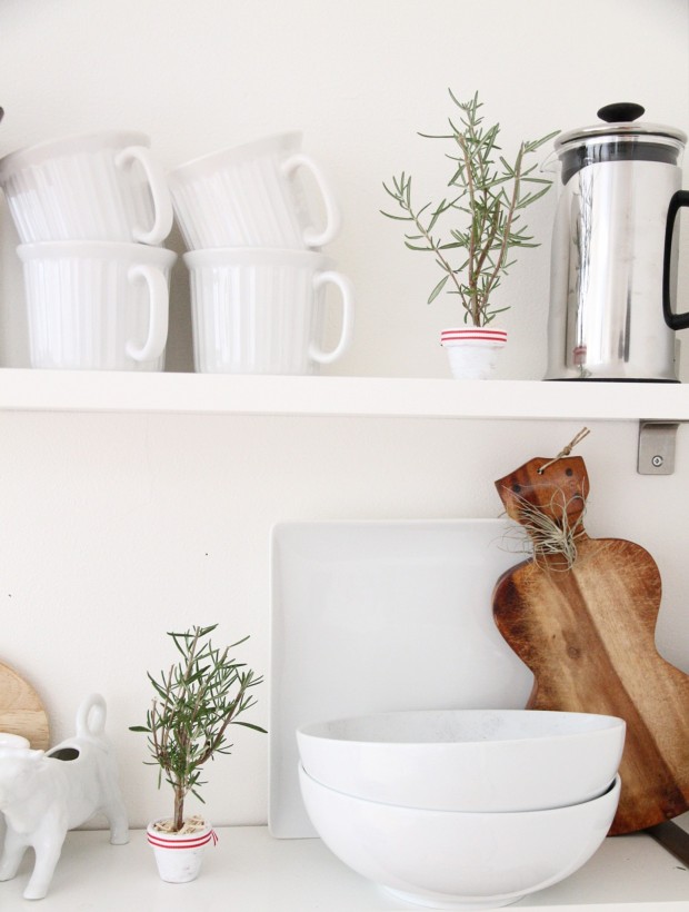 White kitchen with wooden bread board, and white dishes with the little tree on the counter.