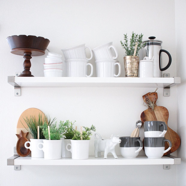 An open shelving unit with cups, cutting boards, a French press and little plants.