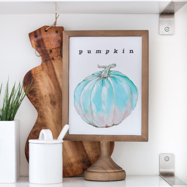 The royal blue pumpkin printable in a wooden frame with a cutting board behind it on the shelf.