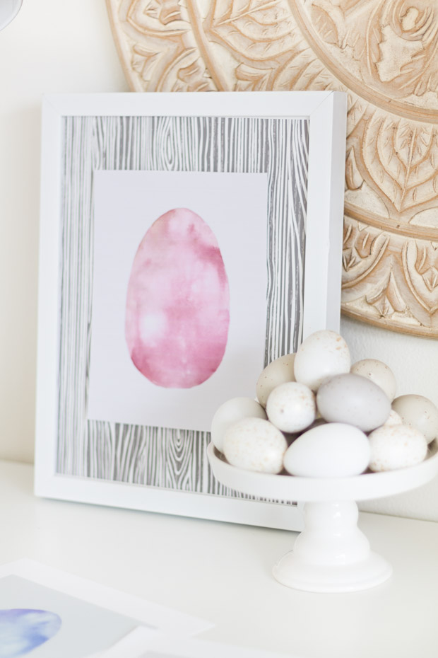 The framed picture beside a cake stand filled with wooden Easter eggs.