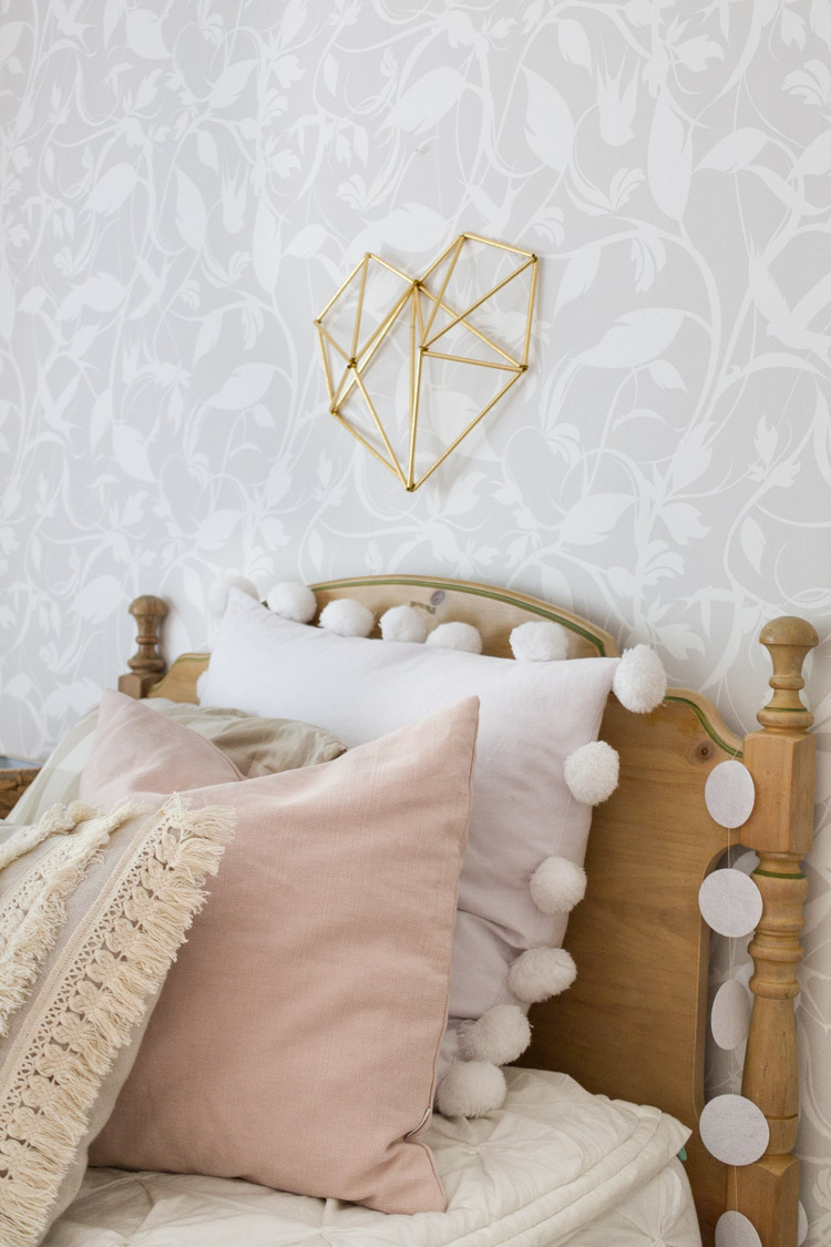 A bed with white and pink pillows, with pom poms on the pillows.   A heart is on the wall just about the headboard.