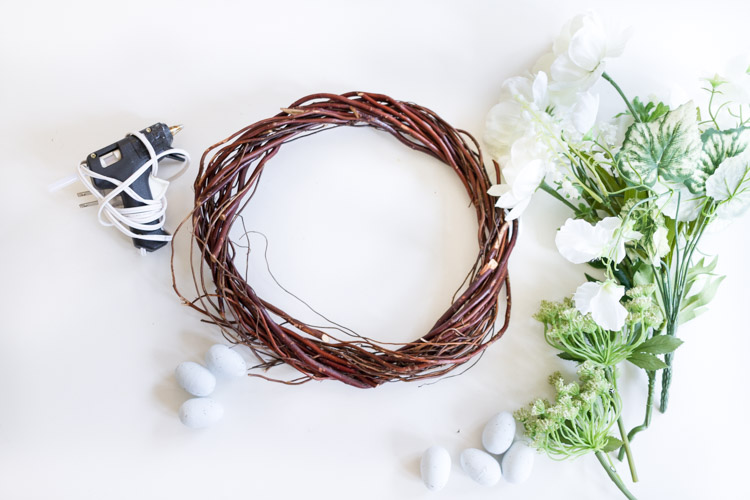 A hot glue gun, wreath, white flowers and stems and miniature faux eggs on the table.