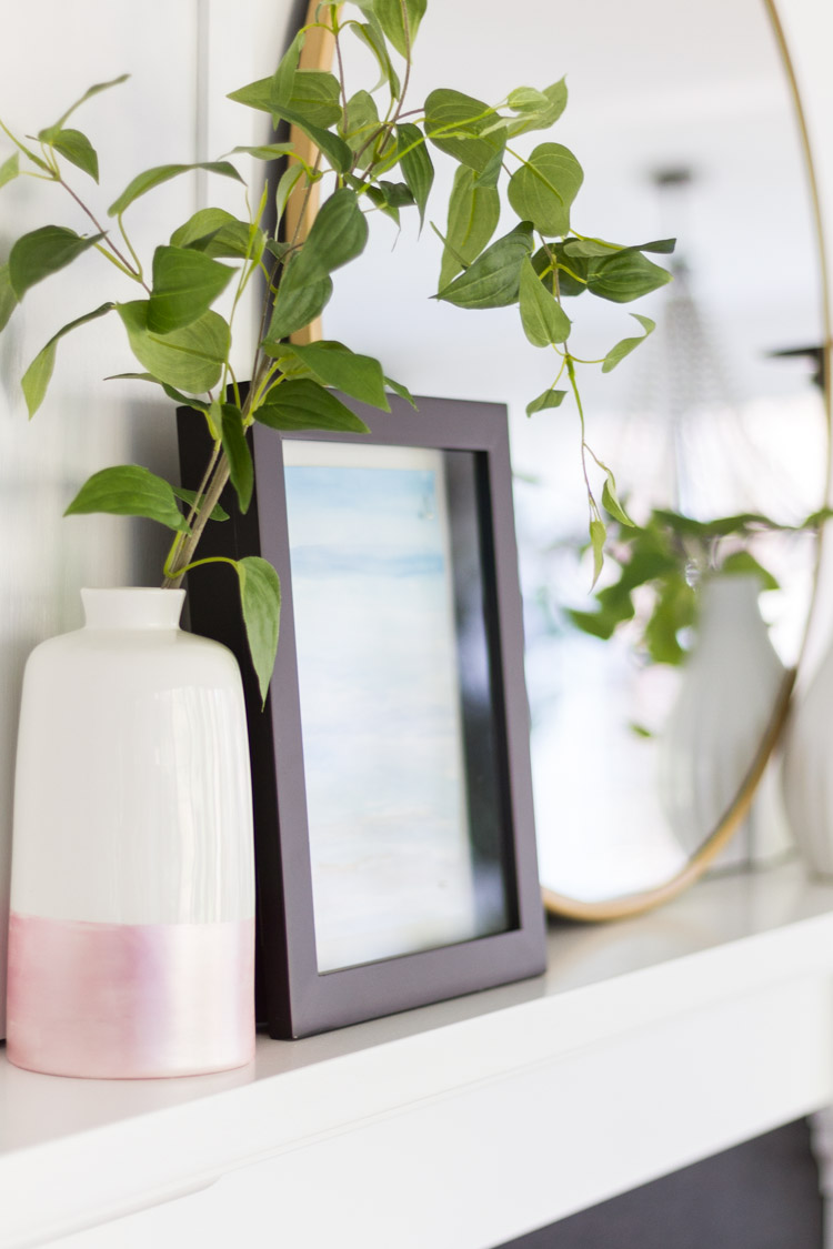 A white and pink vase on the mantel with a branch and green leaves in it.