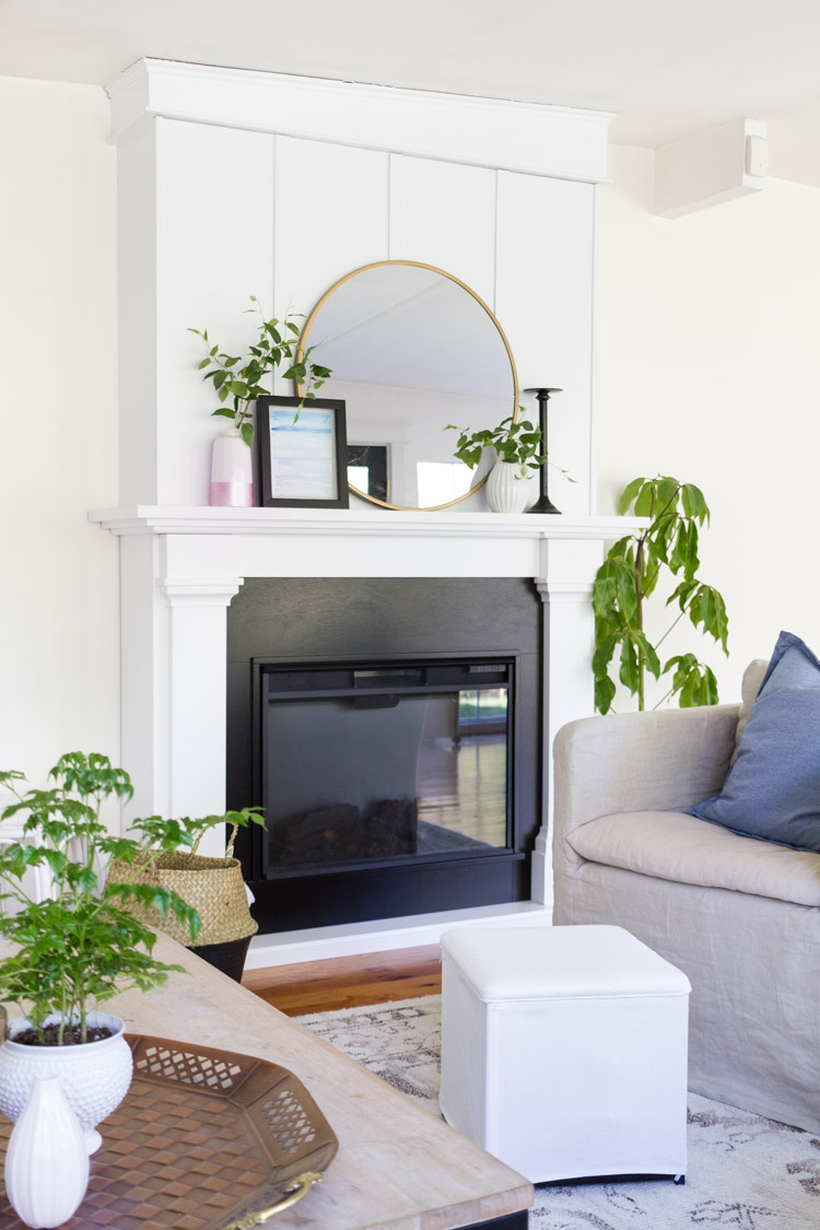 A white living room with green plants, a mirror on the mantel, and a fireplace.
