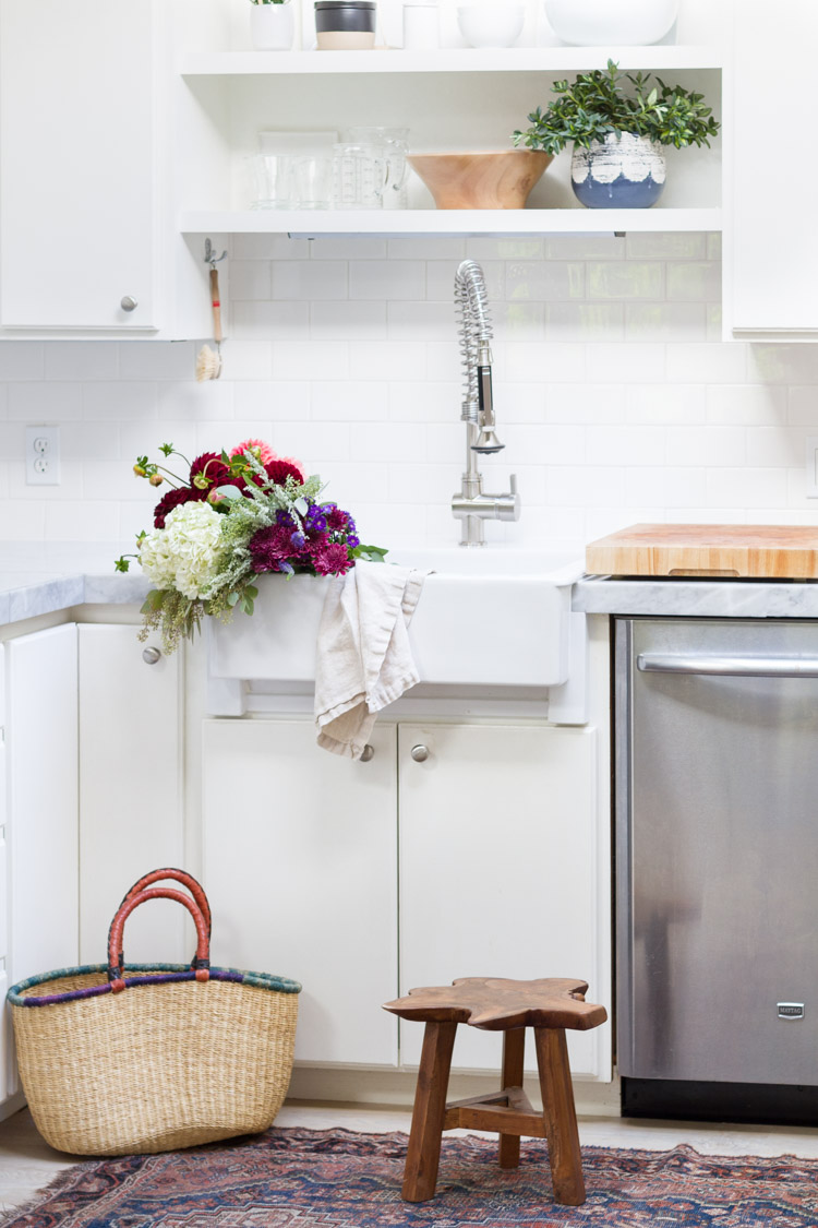 A farmhouse sink with flowers in it, a shelf above it, a wooden step stool and a basket in the kitchen.