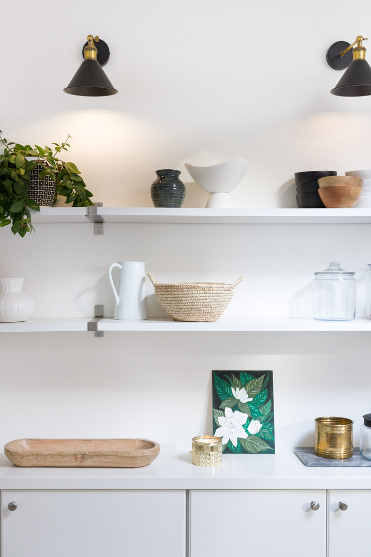 Open shelving in white kitchen with wooden tray, picture, a white pitcher and a small plant.